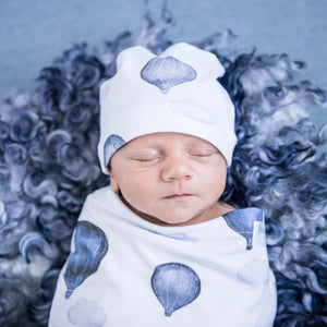 Snuggle Hunny Kids Swaddle and Beanie Set in Cloud Chaser design worn by a sleeping baby on a blue grey wooly rug