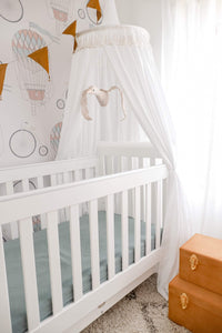 White round canopy hanging over white cot bed in nursery with mustard suitcase storage and mustard bunting and balloon decals on wall