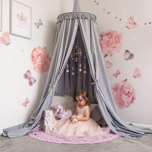Grey Round Canopy over reading nook with large rose decals on wall and girl in tulle dress sitting on cushions underneath with a purple crochet rug on floor. 