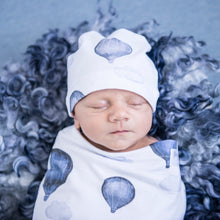 Load image into Gallery viewer, Snuggle Hunny Kids Swaddle and Beanie Set in Cloud Chaser design worn by a sleeping baby on a blue grey wooly rug