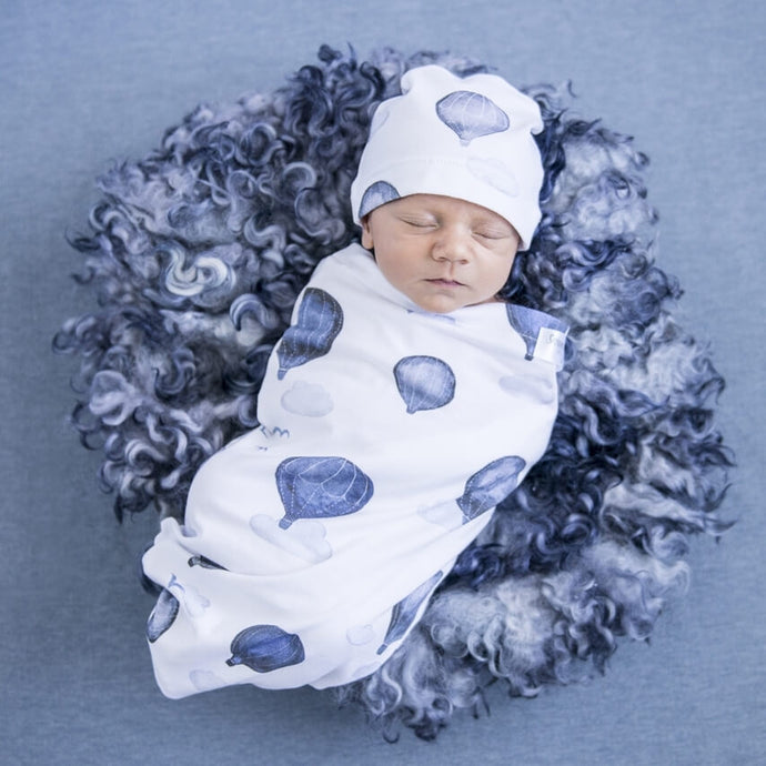 Snuggle Hunny Kids Swaddle and Beanie Set in Cloud Chaser design worn by a sleeping baby on a blue grey wooly rug