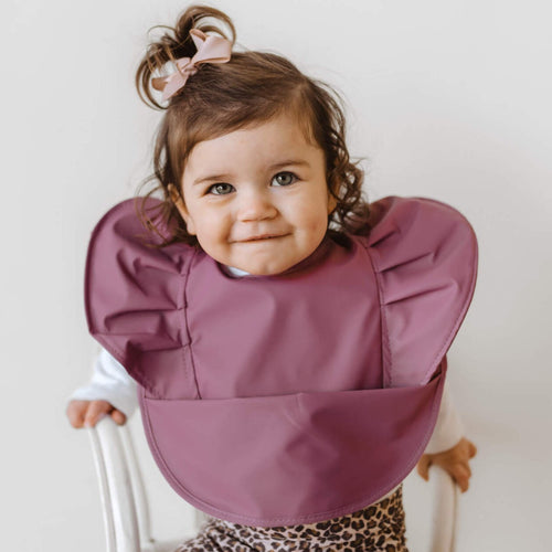 Snuggle Hunny Kids Waterproof Bib in Mauve Frill worn by a toddler with a bow in their hair