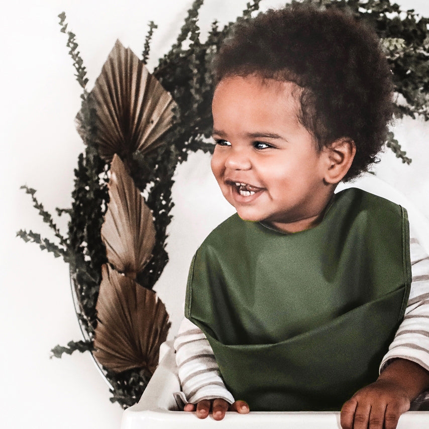 Snuggle Hunny Kids Waterproof Bib in Olive worn by a toddler smiling sitting in a highchair with dried leaves behind