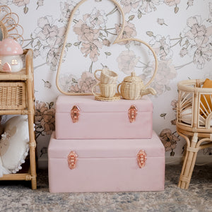 two dusty pink storage cases with rose gold handles sitting on the floor of a girls bedroom with a rattan tea set on top