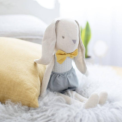 linen soft toy rabbit sitting on bed with mustard bow and grey linen shorts