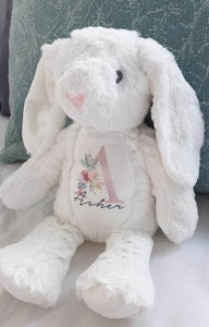 White soft bunny rabbit sitting on bed with long white floppy ears. The name Asher and the Initial A is printed on the bunnys chest with a variety of colourful florals. 