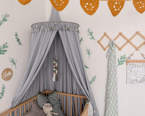 Nursery with grey round canopy over wooden cot and crochet mustard bunting with green leave decals on wall