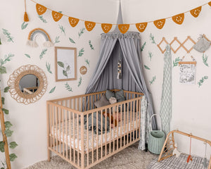 Nursery with grey round canopy over wooden cot and crochet mustard bunting with green leave decals on wall and rattan toys and mirror