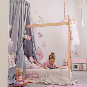 Grey round canopy over timber frame bed with girl lying on bed reading and rose decals on wall with toys around the room and pink accents