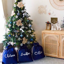Load image into Gallery viewer, Three navy blue personalised santa sacks are sitting under a Christmas tree. The tree is decorated with gold and navy blue decorations. The words Merry Christmas are on the wall beside the tree and there is a wicker cabinet sitting next to the tree. 