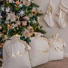Load image into Gallery viewer, three ivory santa sacks sitting under a green chsitmas tree personalised with names in gold metallic font. 