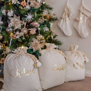 three ivory santa sacks sitting under a green chsitmas tree personalised with names in gold metallic font. 