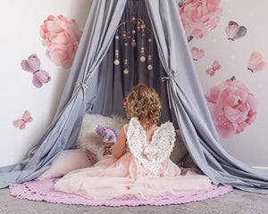 Grey Round Canopy over play nook with large rose decals on wall and a girl in tulle dress with lace fairy wings sitting on cushions underneath with a purple crochet rug on floor.