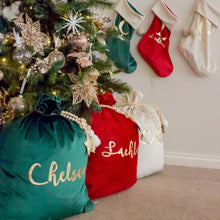 Load image into Gallery viewer, green, red and ivory santa sacks sitting under a christmas tree with matching stockings hanging on the wall beside the tree.