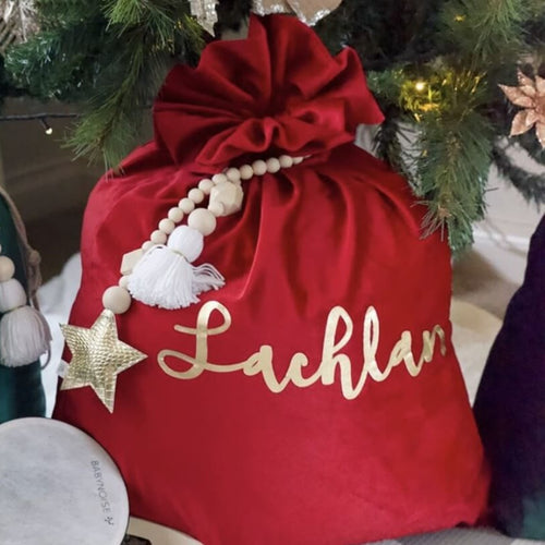 red velvet santa sack with gold writing name lachlan on the front of the sack. A beaded gold garland is hanging around the top of the sack and the sack is sitting under a green christmas tree