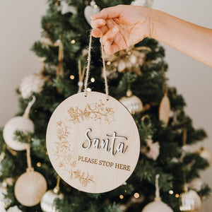 ladies hand holding a round timber sign that reads santa please stop here. A christmas tree with gold and silver decorations is visible in the background.