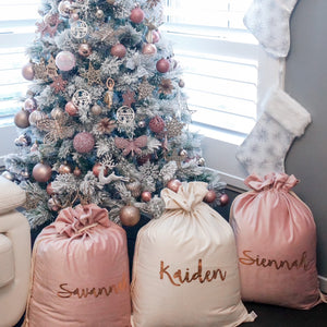 Christmas tree decorated with pink, white and rose gold decorations. Two pink and one ivory santa sacks are sitting in front of the tree and two white and silver stockings are hanging on the wall above the sacks. 