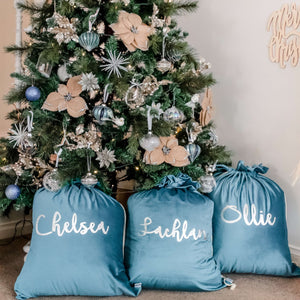 Three turquoise santa sacks sitting under a christmas tree personalised with names in silver writing.