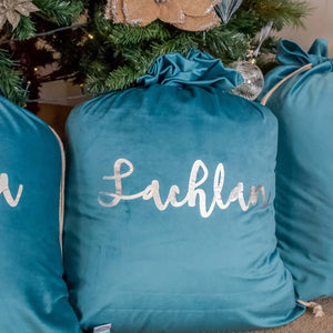 three turquoise blue santa sacks sitting under a christmas tree. The middle sack in personalised with the name Lachlan written in silver metallic font. 