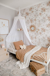 White round canopy over white wrought iron bed with featured wall of rose wallpaper and dusty peach bedspread and pillow and rattan mirror and chest