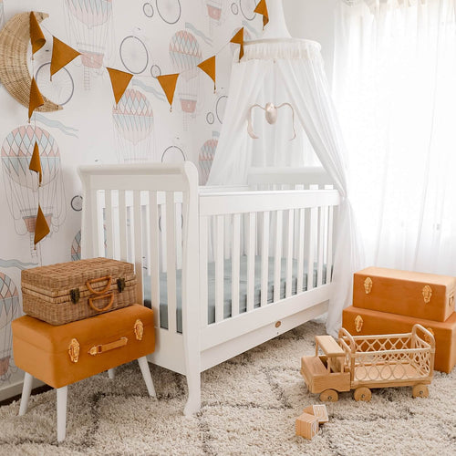 White round canopy hanging over white cot bed in nursery with mustard suitcase storage and mustard bunting with rattan toy truck on floor