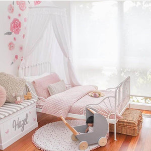 Girls room with white round canopy hanging over wrought iron bed with rose decals on wall and pink quilted bedspread with wooden pram on floor